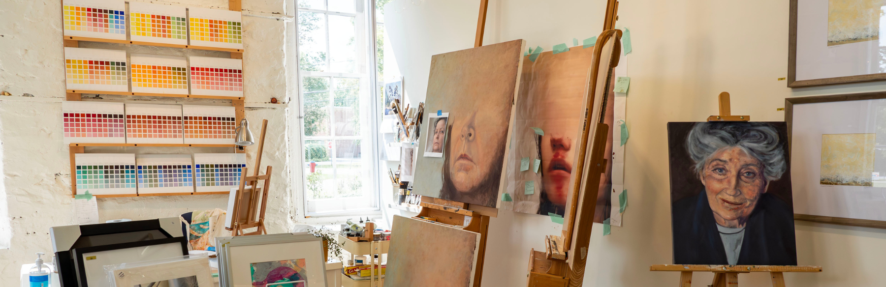 Image of an artists studio with canvas on easels to the left side of the image. The canvas are half painted images of faces. On the back wall there are a bunch of colour scale images on a shelf 