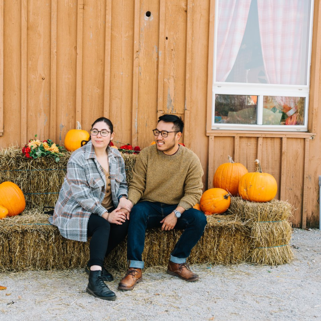 A couple holding hands sitting on bales of hay with some pumpkins surrounding them 