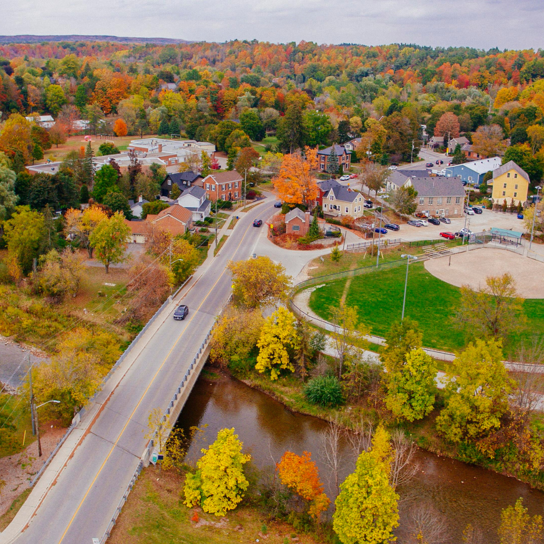 An aerial image of Downtown Glen Williams. You can see the credit river passing under a bridge. On the right side of the photo you can see a baseball diamond and behind the diamond there is a yellow building, blue building and some smaller brown buildings to the left. 