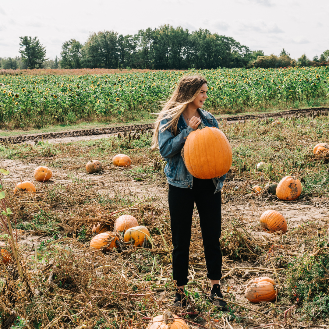 An image of a young woman standing in the middle of a pumpkin patch. She is holding a big orange pumpkin in her hands