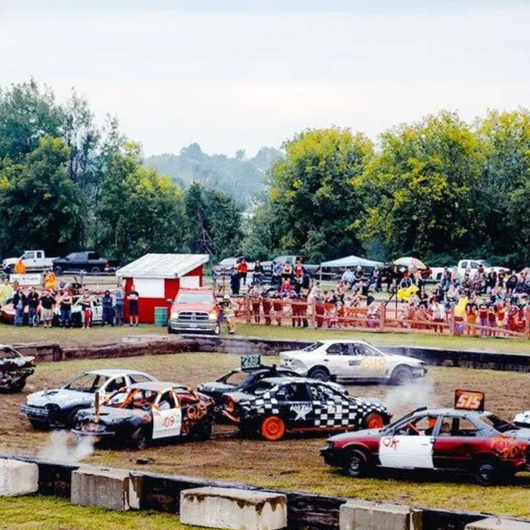 A bunch of cars participating in a car derby. A bunch of them are all smashed into each other and a couple of cars are smoking from their hoods 