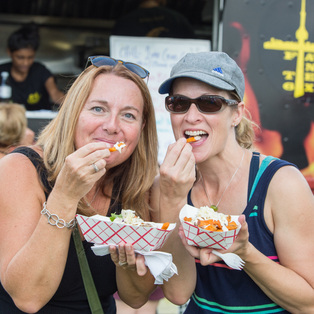 Two women standing in front of a good truck eating loaded french fries 