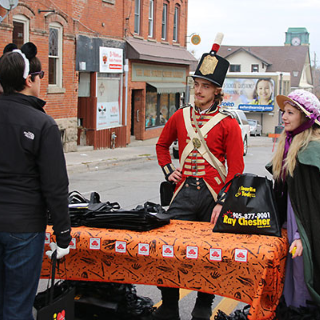 two people standing behind another table talking to another man. The two people behind the table are dressed as a solider and an old timey woman 