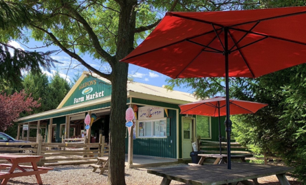 A picture of a green barn with a sign on it that says Allison's Farm Market. In the foreground there is some picnic benches with red umbrellas 