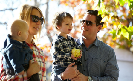 a mom and dad both holding two young children. the father is holding a little boy who has a apple in his hand